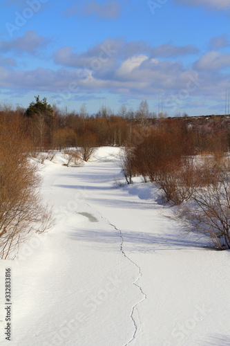 Fototapeta Naklejka Na Ścianę i Meble -  snowy road between shrubs on a background of a winter forest and blue bright sky with white clouds. Pastoral idealistic landscape, winter magical mood. Stock photo for web and print.