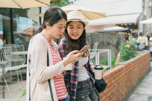 Image of cheerful asian korean women travelers in casual clothes drinking takeaway coffee and using smartphone on red brick wall outdoors. two ladies enjoy sunshine sharing mobile phone together. © PR Image Factory
