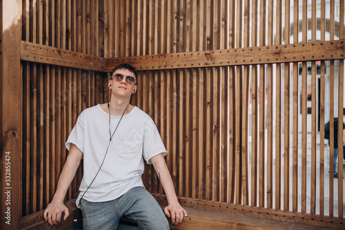 a young guy in dark glasses, a white t-shirt and blue jeans walks along the street and listens to music with headphones