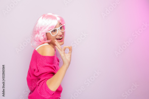 Caucasian woman with pink wig and sweater, wearing white glasses, posing funny at the photo studio. Funny expressive women and colorful portrait concept. Pink.