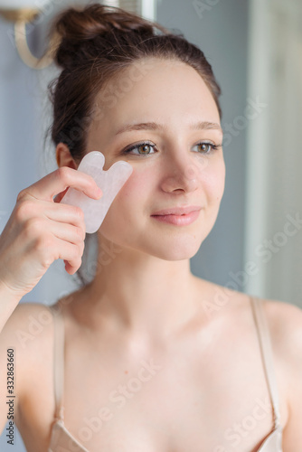 The girl makes a massage gua sha in light bathroom in home. Rose quartz massager, natural stone. Lifting, natural care.