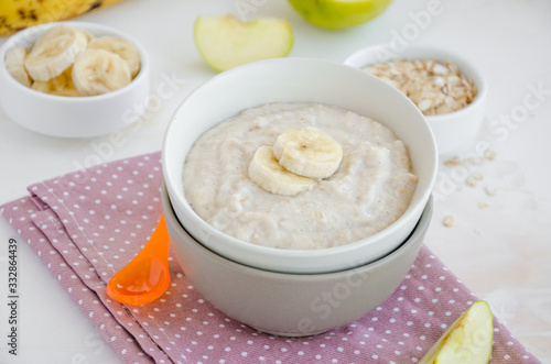 Baby food. Creamy oatmeal with banana slices and apple in a bowl with a spoon on a light background. Healthy breakfast. Porridge for breakfast. Horizontal orientation. Close up.