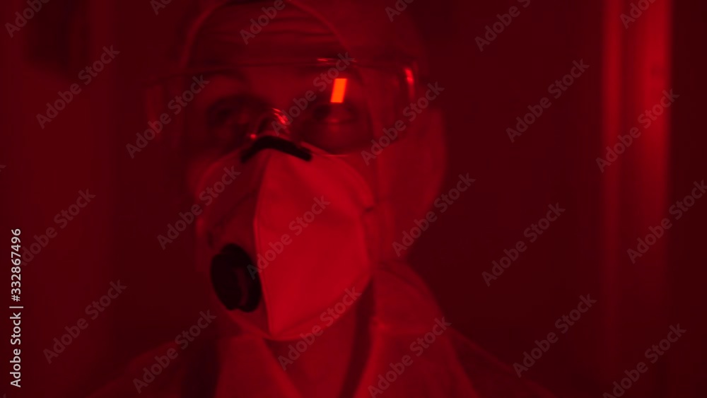 Man wears protective suit, face mask and goggles, looks anxiously around dark misty room with flashing red blue light; close up Man wears protective suit, face mask and goggles, looks anxiously around dark misty room with flashing red blue light; close up