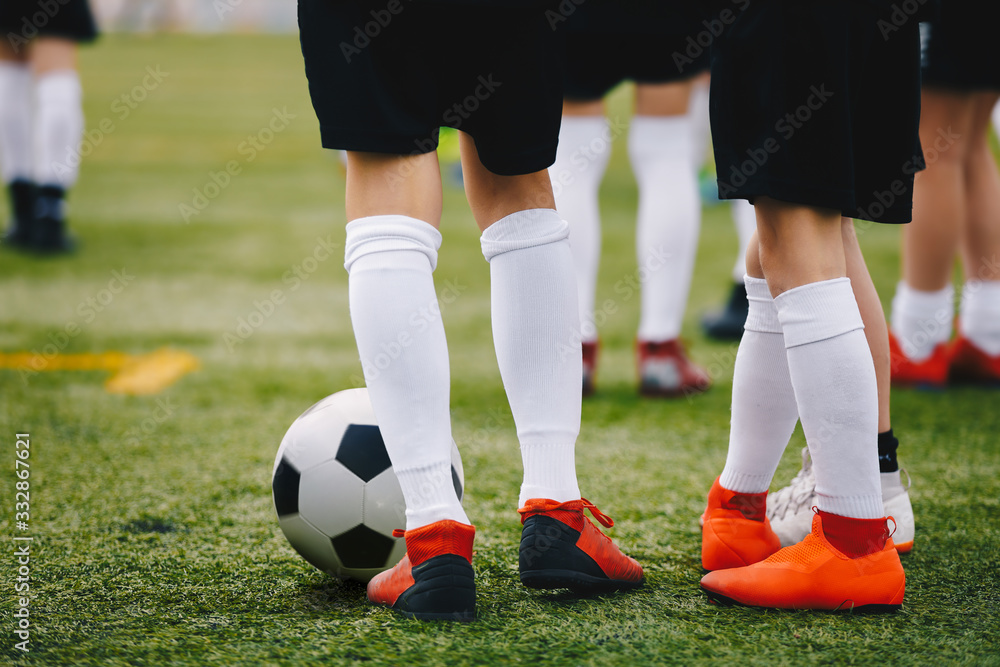 Fototapeta premium Soccer players with ball on a training field. Young junior level athletes in football cleats and uniforms on grass training ground. Closeup image of soccer players legs. Football background