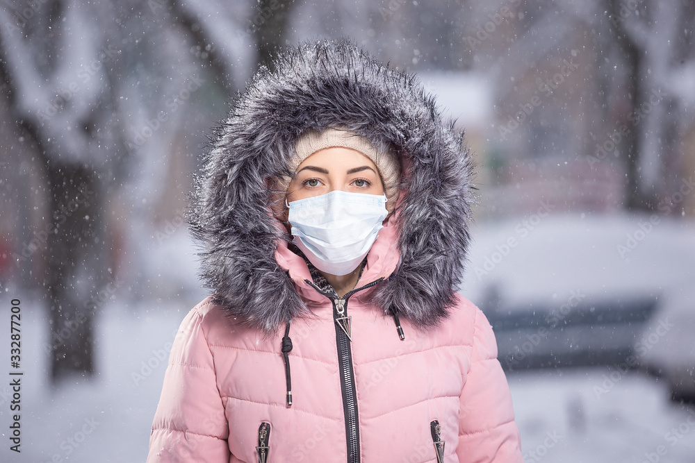 Portrait of woman in pink walking down the street in winter under snowfall in a protective mask to protect against infectious diseases. Protection against colds, flu, air pollution. Health concept