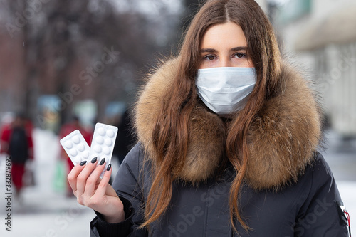 Portrait of a beautiful girl in a medical protective mask in holding white pills in her hands for colds and flu. Winter street portrait of a woman under snowfall. © simikov