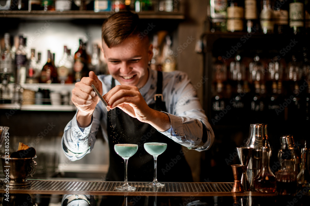 young smiling barman decorates two glasses with cocktails.