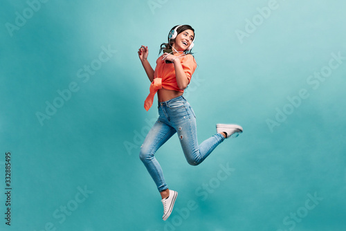 Young beautiful energy girl with white headphones listening to music laughs and jump on blue background in studio and looks away.Dressed in an orange shirt and light jeans, holding a phone. photo