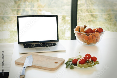 Laptop with blank white screen and vegetables, knife and fruits on countertop in modern kitchen with big windows, copy space. Culinary courses or recipe online. Cooking home concept. Food blog