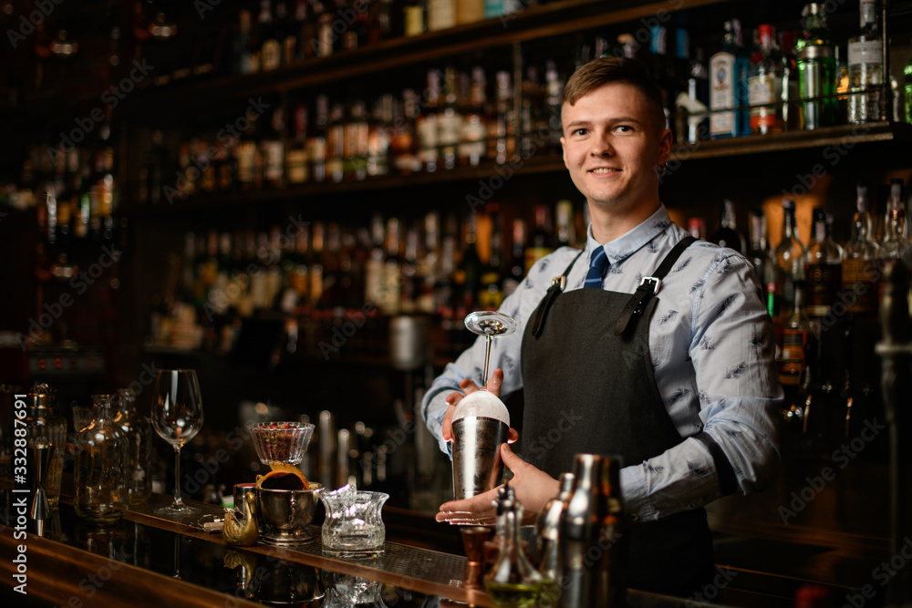 Professional young smiling barman preparing glass and shaker to make cocktail.
