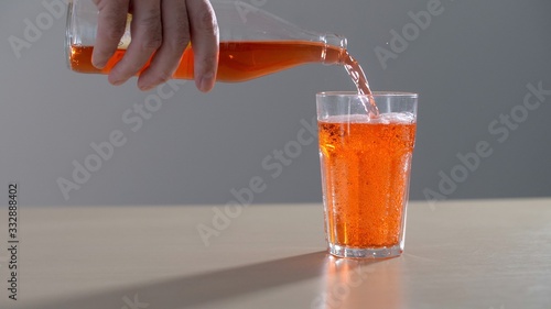 Hand pouring an orange carbonated drink from glass bottle into a glass. Male hand Holding orange soda bottle tight and close pours Into a glass Outside. Pouring fresh and cold yellow lemonade drink.