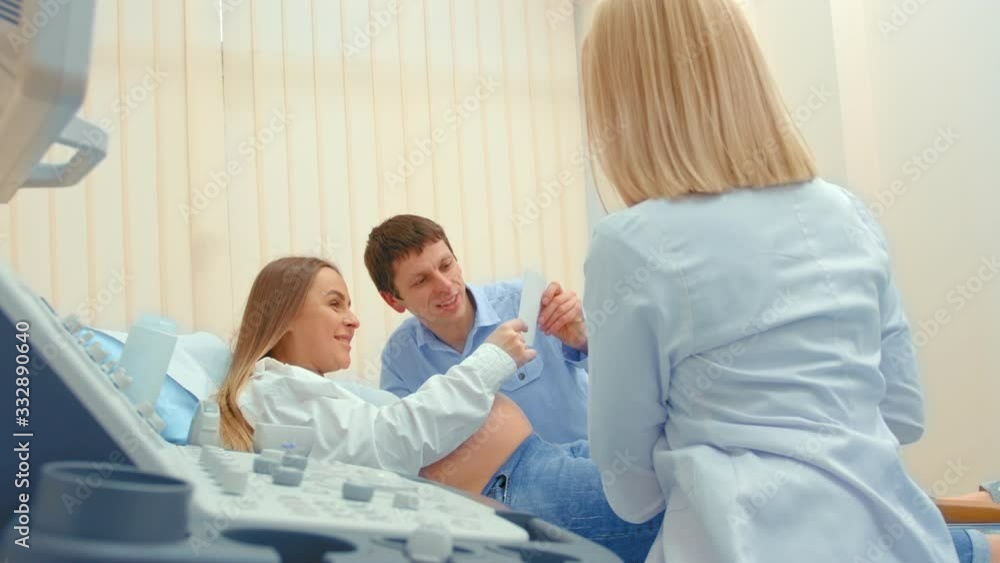 A young woman doctor shows a photo of an ultrasound scan of a child. A pregnant woman rests on a couch, a young couple happy and smiling
