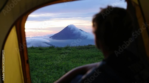 Cinematic close up of caucasian man sitting inside a yellow tent looking out into the distance towards the smoking Momotombo volcano in Nicaragua during sunrise. photo