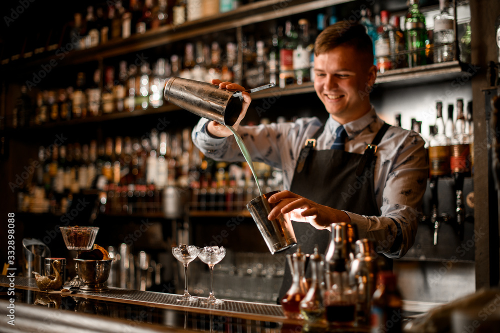 young smiling barman pours cocktail in shaker. Two glasses with ice stand on bar.