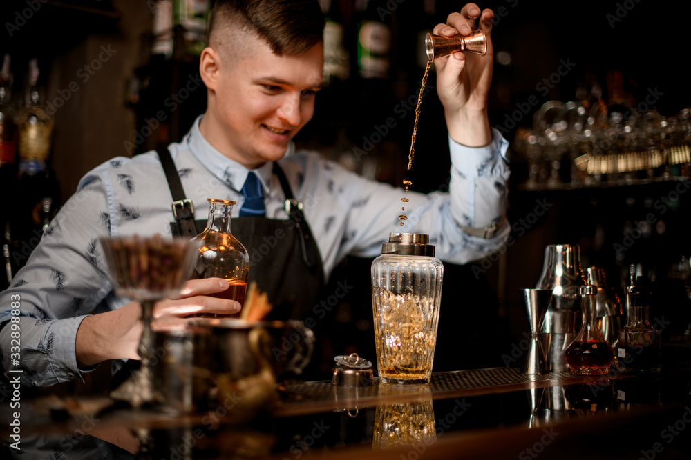 young smiling bartender at bar pours drink into shaker using beaker