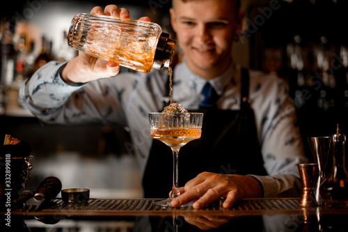 young smiling bartender professionally pours drink from shaker into glass.
