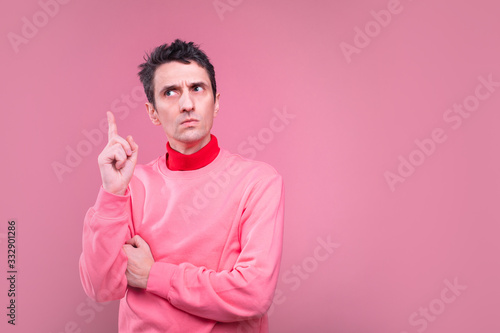 Serius concentrated young man in pink sweater point up with smart face. Stand alone in studio and pose. Isolated over pink background.