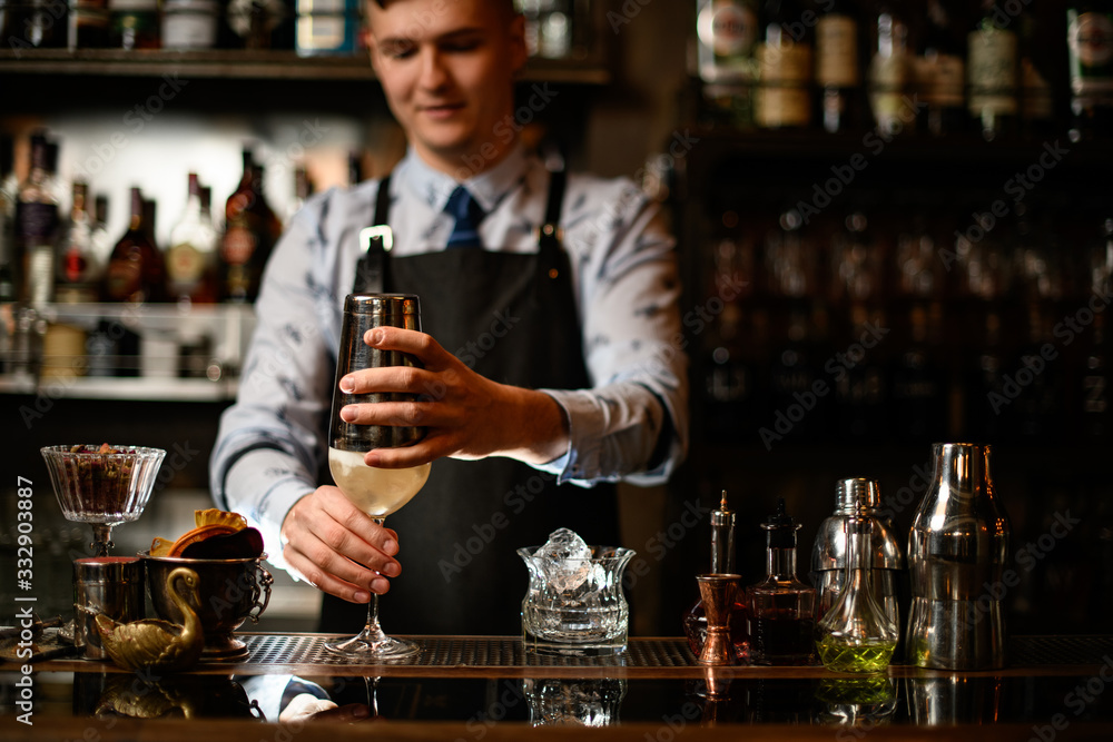 Young barman holds shaker by hand over glass with cocktail.