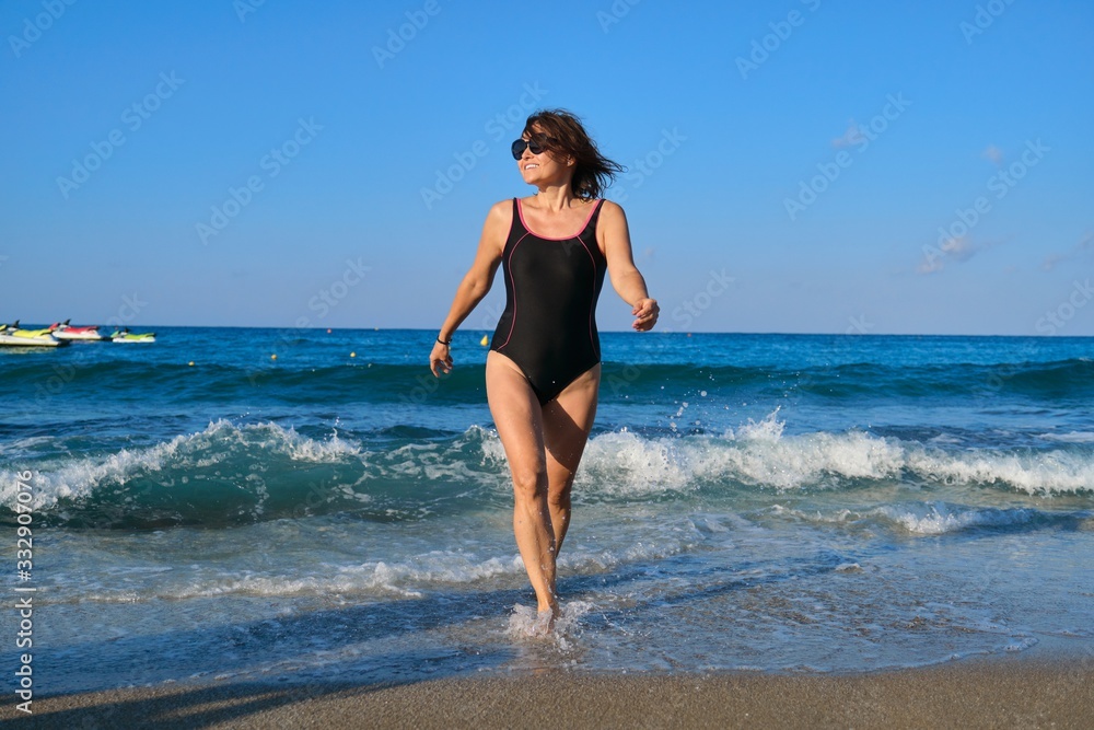 Mature smiling woman in swimsuit with sunglasses walking along beach