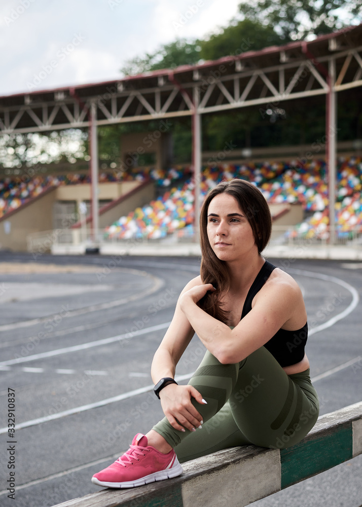 Young brunette woman, wearing black top and green leggings, sitting on city stadium on summer morning, resting relaxing after training. Portrait of sporty girl, posing. Healthy active life concept