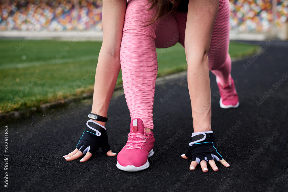Young woman, wearing pink leggings, sneakers, black top and fitness gloves, training on city stadium on summer morning. Sportswoman, stretching, preparing to run outside. Healthy lifestyle concept.