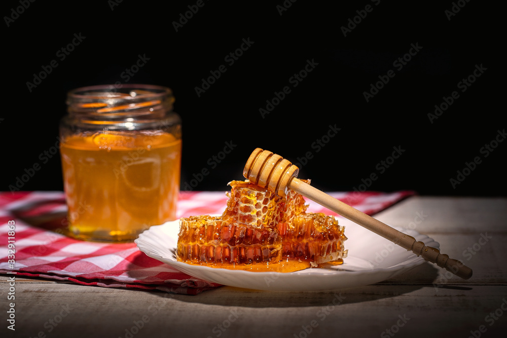 glass jar full of honey, plate with honeycomb and dipper on wooden table with checkered tablecloth and dark background glass jar full of honey, plate with honeycomb and dipper on wooden table with checkered tablecloth and dark background