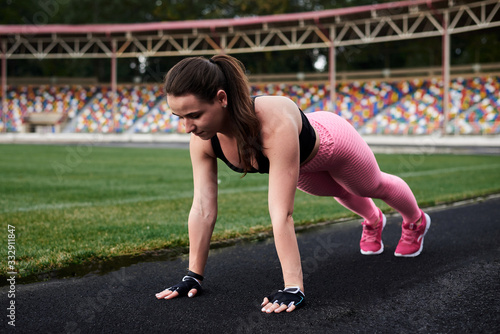 Young brunette woman  wearing pink leggings  sneakers  black top and fitness gloves  training on city stadium in morning. Sportswoman  doing plank on the ground outside. Healthy lifestyle concept.