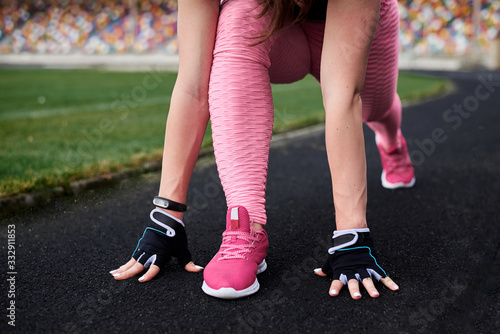 Young woman, wearing pink leggings, sneakers, black top and fitness gloves, training on city stadium on summer morning. Sportswoman, stretching, preparing to run outside. Healthy lifestyle concept.