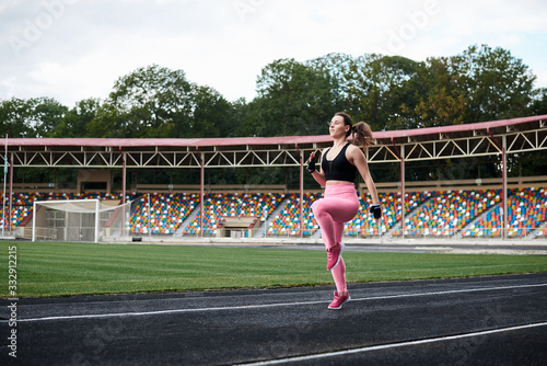 Young brunette woman, wearing pink leggings, sneakers and black top, training on city stadium on summer morning. Sportswoman, running jogging outside to loose weight. Healthy lifestyle concept.