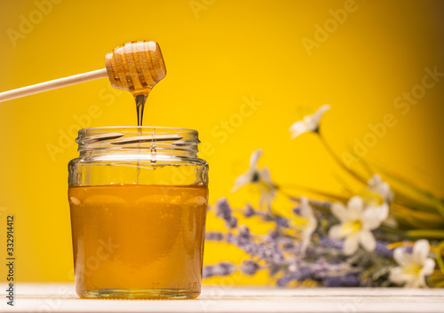 glass jar full of honey and dipper on wooden table with flowers on background