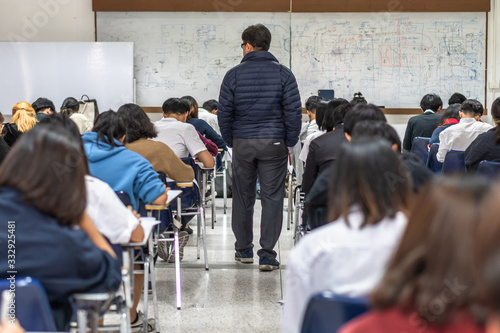 School exam room with teacher (invigilator) monitoring students taking educational examination in classroom for admission test photo