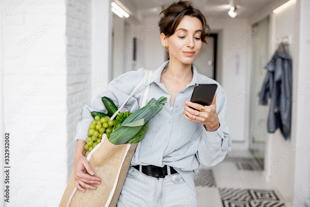 Portrait of a young and cheerful woman standing with mobile phone and shopping bag full of fresh food at home. Concept of buying products online Portrait of a young and cheerful woman standing with mobile phone and shopping bag full of fresh food at home. Concept of buying products online