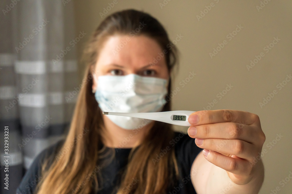 A girl in a medical mask holds a thermometer in her hand, which shows a high value of body temperature. Symptoms of coronavirus. Quarantine, work from home.