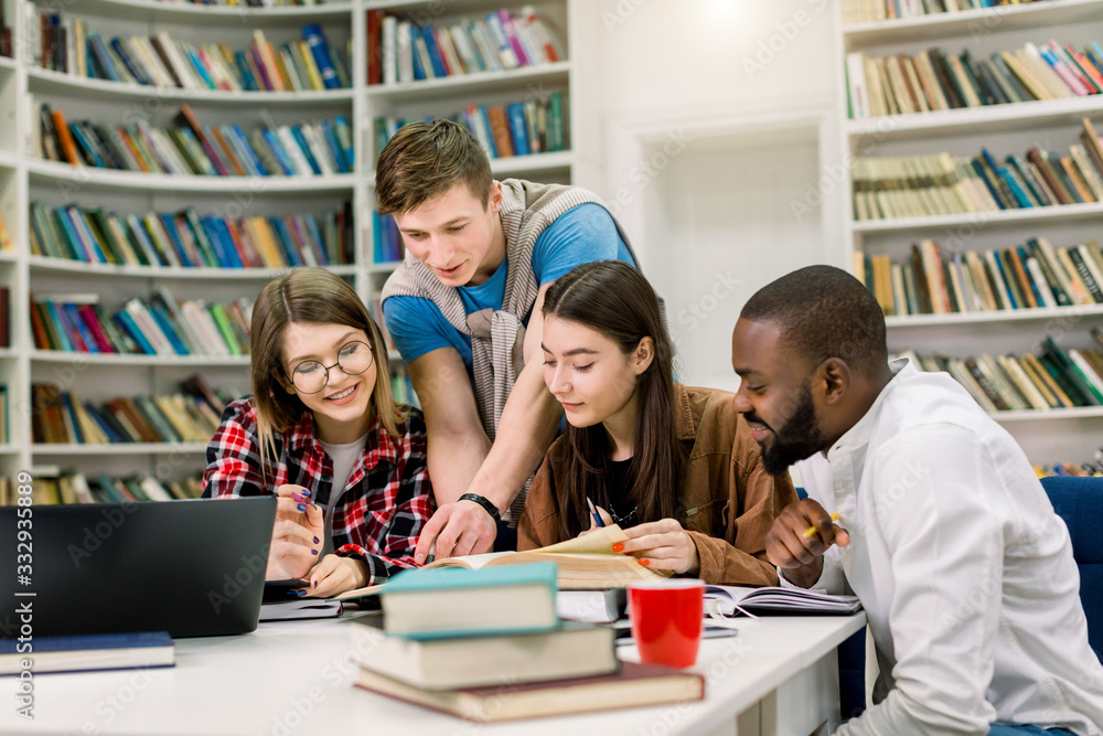 Four positive smiling multiethnic boys and girls sitting at the table in modern college library and jointly doing home task or searching information for learning subject Four positive smiling multiethnic boys and girls sitting at the table in modern college library and jointly doing home task or searching information for learning subject
