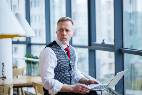 Adult male businessman, teacher, mentor working on a new project. Sits by a large window on the table. He looks at the laptop screen.