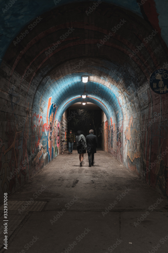A couple walking through a colorful tunnel at night. lights are illuminating the pathway as a man walks towards them being busy with his phone. The people are slightly blurred to show their movement. 