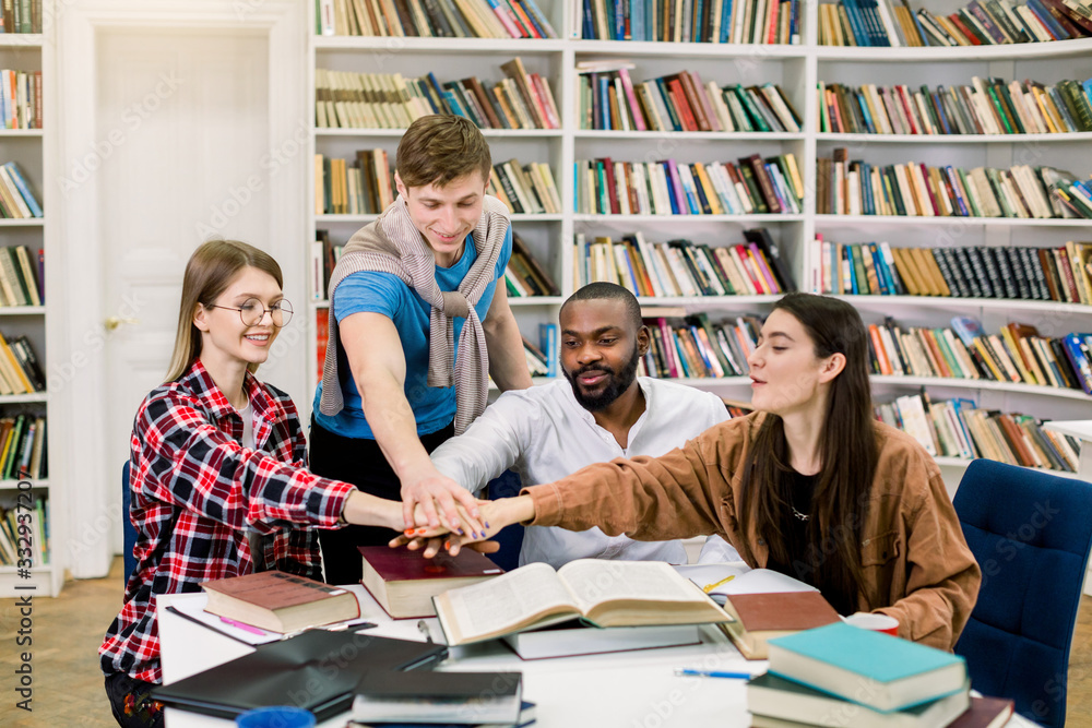 Group of young joyful multiracial college students collaborating on project, sitting at the table in library and putting hands together Group of young joyful multiracial college students collaborating on project, sitting at the table in library and putting hands together