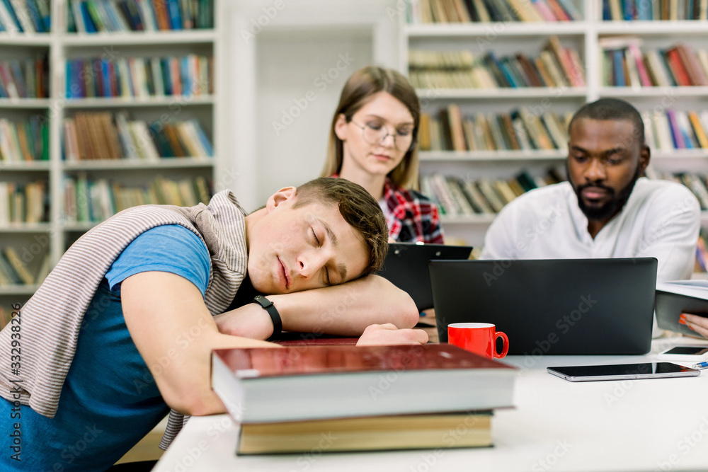 Close up of tired sleeping handsome male student lying on his hands and books on table. Concentrated attantive multiethnic students preparing for exams, sitting at the table on the background Close up of tired sleeping handsome male student lying on his hands and books on table. Concentrated attantive multiethnic students preparing for exams, sitting at the table on the background