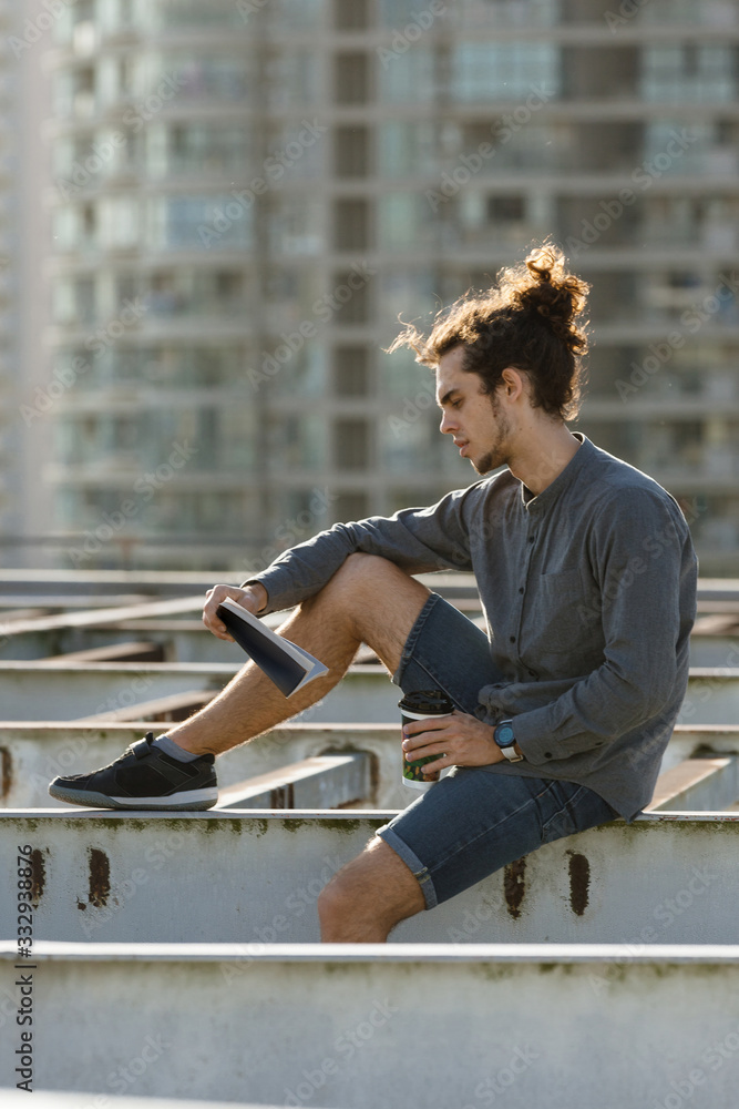 Man sitting on roof and reading book, drinking coffee, tea, drink in paper cup in grey shirt, blue jeans shorts, curly hair collected in high ponytale . Background cityscapes. Urban lifestyle concept