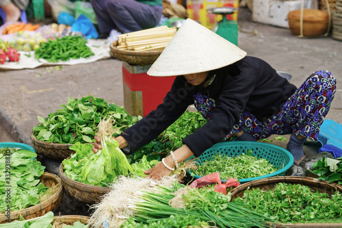 Asian trader in a traditional hat selling fresh greens in the street market photo