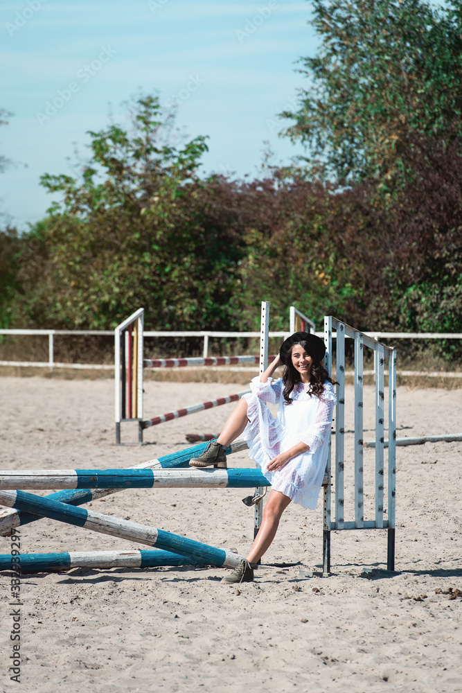 Fototapeta premium Beautiful sexy girl posing on the horizontal crosspiece on a horse epodrom. Female wearing white dress, black hat and black boots