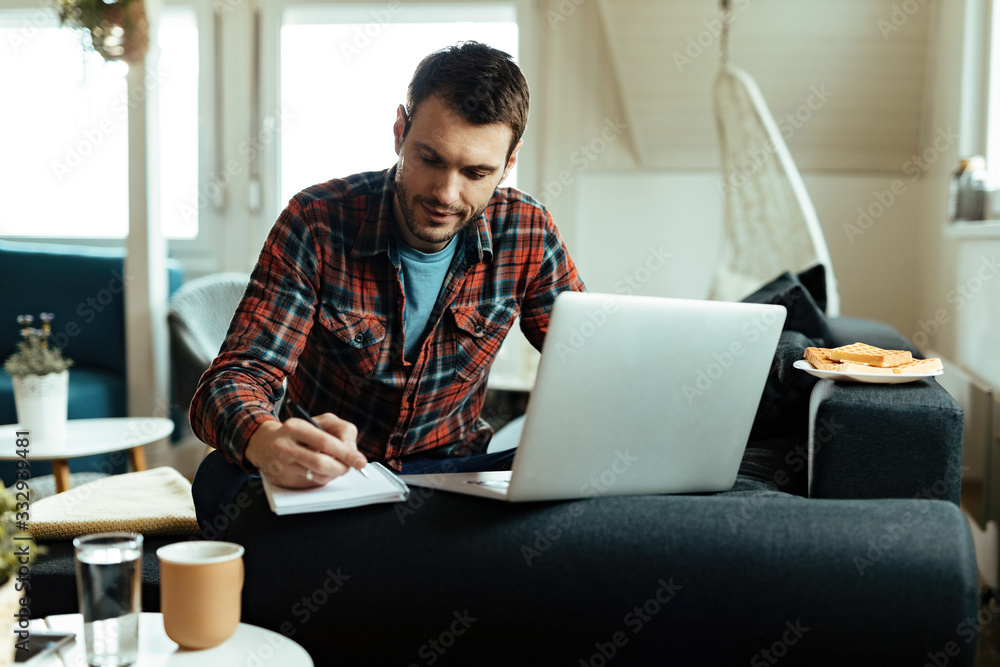 © Drazen - Smiling man writing notes while using computer in the living room. © Drazen - Smiling man writing notes while using computer in the living room.