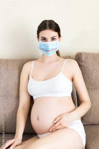 Sick pregnant woman is sitting on the couch at home. Protect your health with protective medical mask. Coronavirus infection. Global quarantine