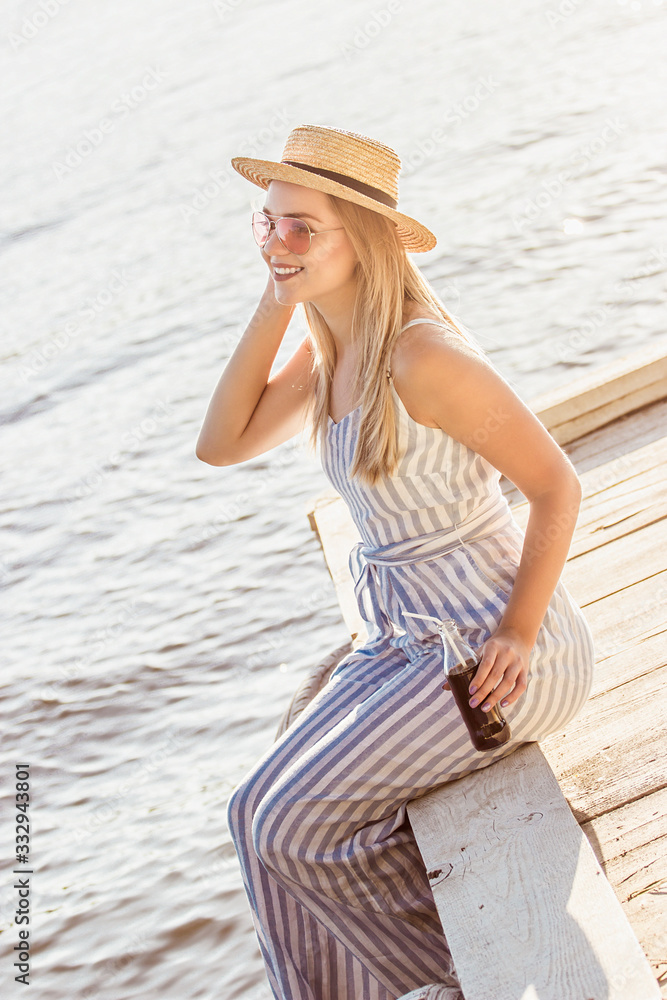 Tender happy blonde girl in stylish summer clothing sitting on wooden pier and drinking lemonade. Cheerful beautiful young woman in stripped overall and straw hat posing at seaside during sunset