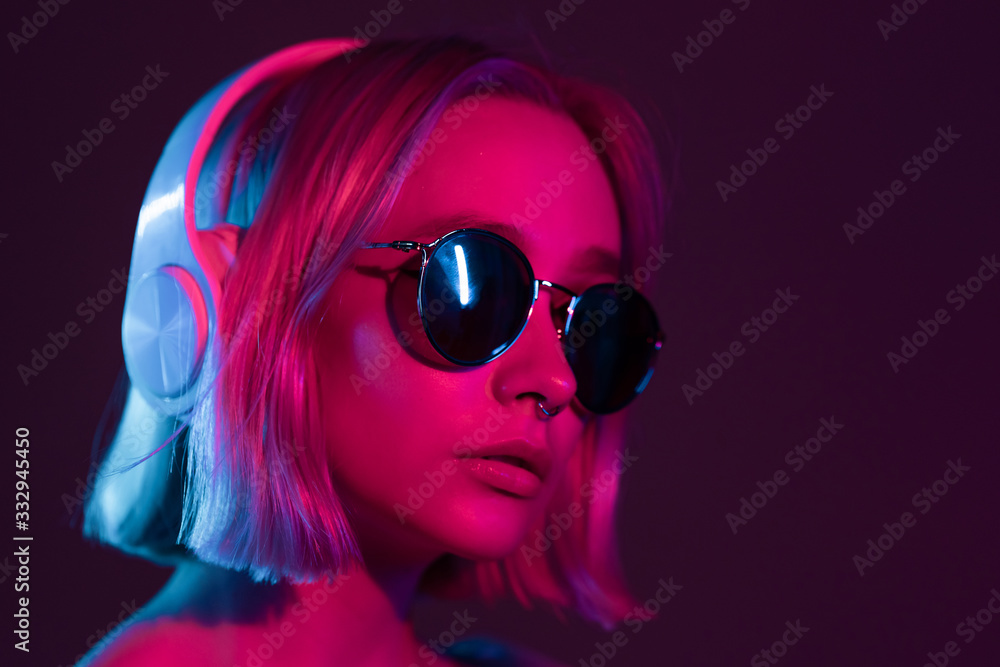 Gorgeous lady with dyed hair and sunglasses listening music in headphones and singing with neon light background. Charming hipster girl dancing with eyes closed.