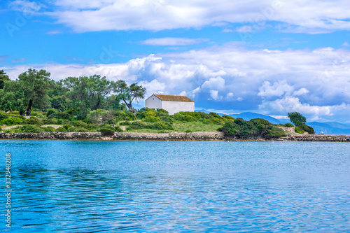 Beautiful summer landscape – calm blue sea water, park with green trees and bushes, old white house and mountains on the horizon. Paxos Island, Greece. 