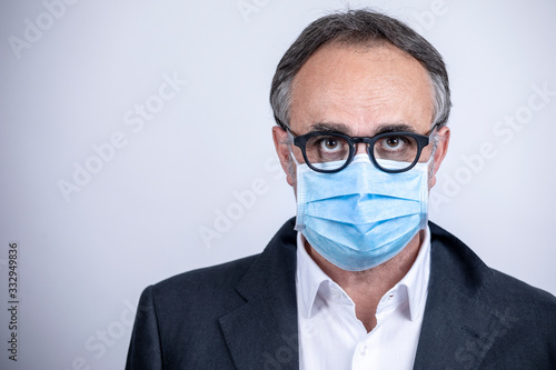 Ritratto di uomo di mezza età con occhiali da vista che indossa una mascherina chirurgica azzurra isolato su sfondo chiaro neutro © alex.pin