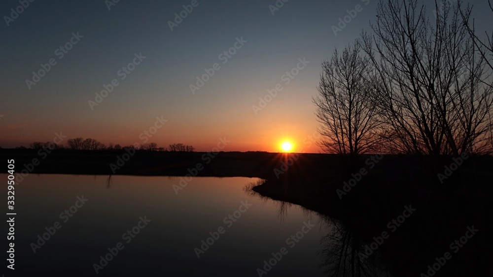 Spring sunset above calm water of artificial fishing pond, with some naked tall broadleaf tree silhouettes on right bank.