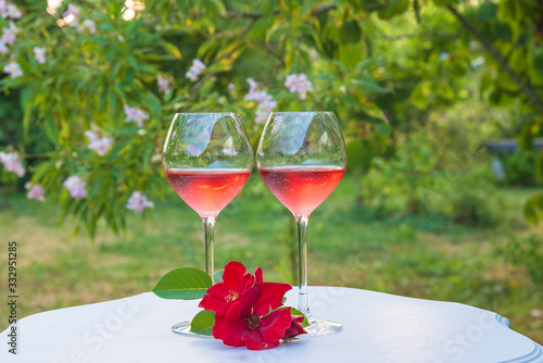 Romantic holiday composition. Two glasses of pink wine and red roses on white vintage table in the blooming garden