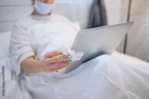 Unrecognizable woman in face mask in bedroom during coronavirus isolation home quarantine cleaning laptop by hand sanitizer, using cotton wool with alcohol to wipe to avoid contaminating with COVID-19